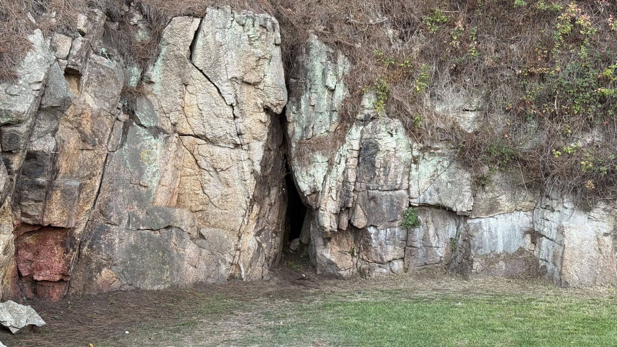 Narrow, dark crevice or small cave entrance between large weathered rock walls covered with patches of moss and dry vegetation.